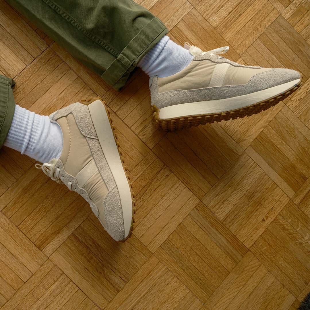 Top-down view of someone's feet wearing the Acorn Trainer in the color Cloud, sitting on a hardwood floor.