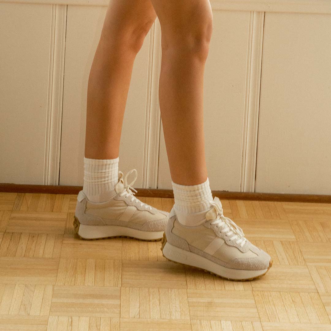Close up view of someone's legs standing on a hardwood flour, wearing the Acorn Trainer in the color Cloud.
