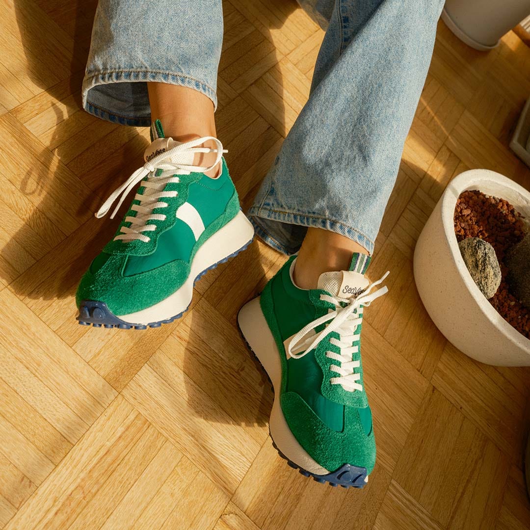 Close up lifestyle shot of a person wearing the Acorn Trainer in Grass Green and sitting on a wooden floor.