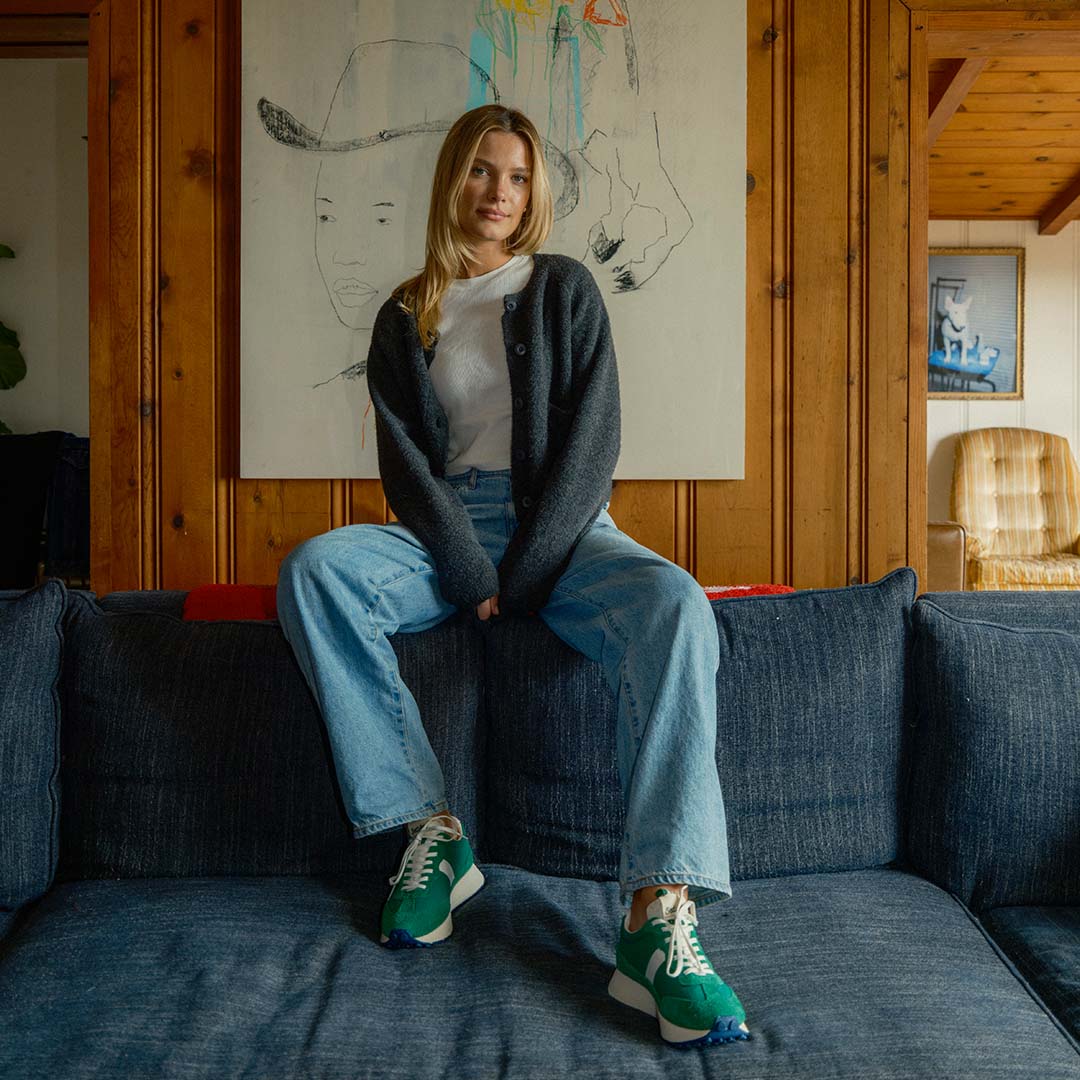 Person sitting on top of a couch, styled in a white tee shirt, dark colored cardigan, blue jeans, and the Acorn Trainer in the color Grass Green.