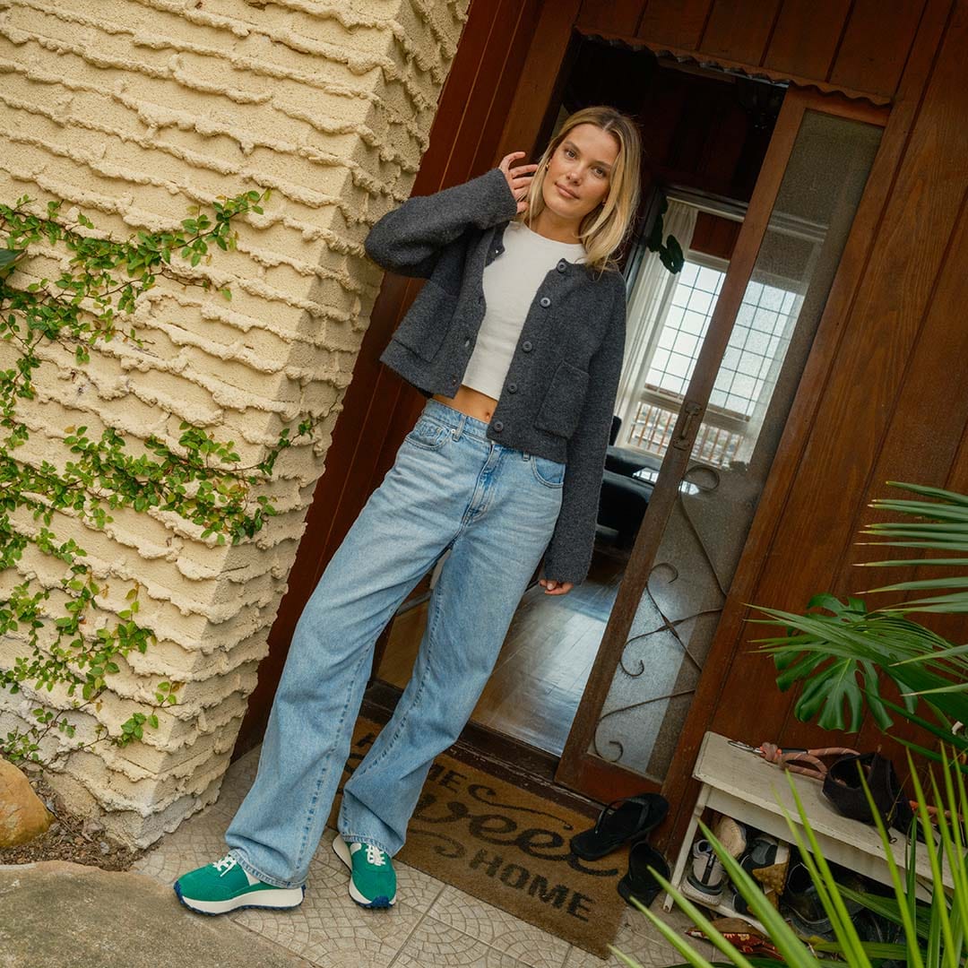 Person standing in the doorway of a house, wearing a gray sweater, white tee shirt, blue jeans, and the Acorn Trainer in the color Grass Green.
