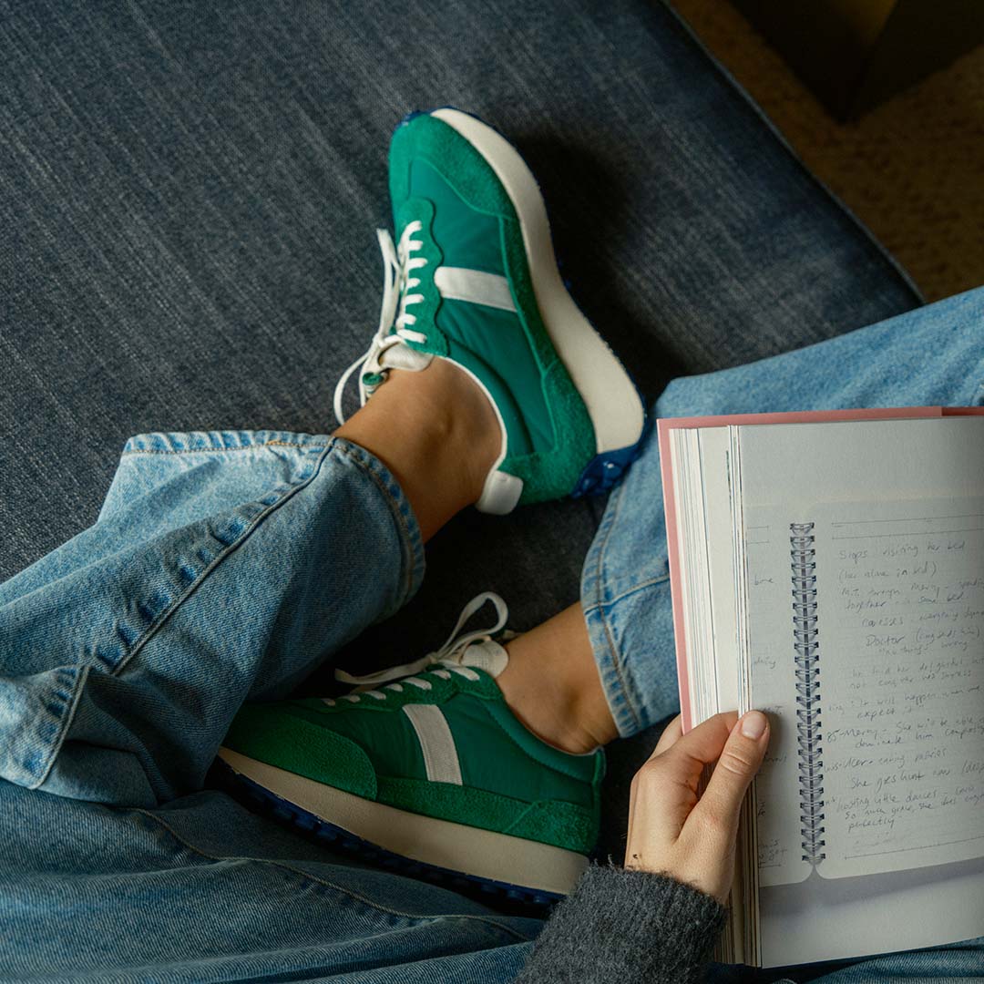 Close up of the Acorn Trainer in the color Grass Green, modeled on a person wearing blue jeans and holding a book.