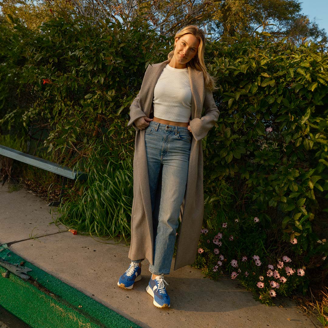 Person wearing the Acorn Trainer in the color Varsity Blue, wearing blue jeans, a white tee shirt, and trench coat, standing against a line of bushes.