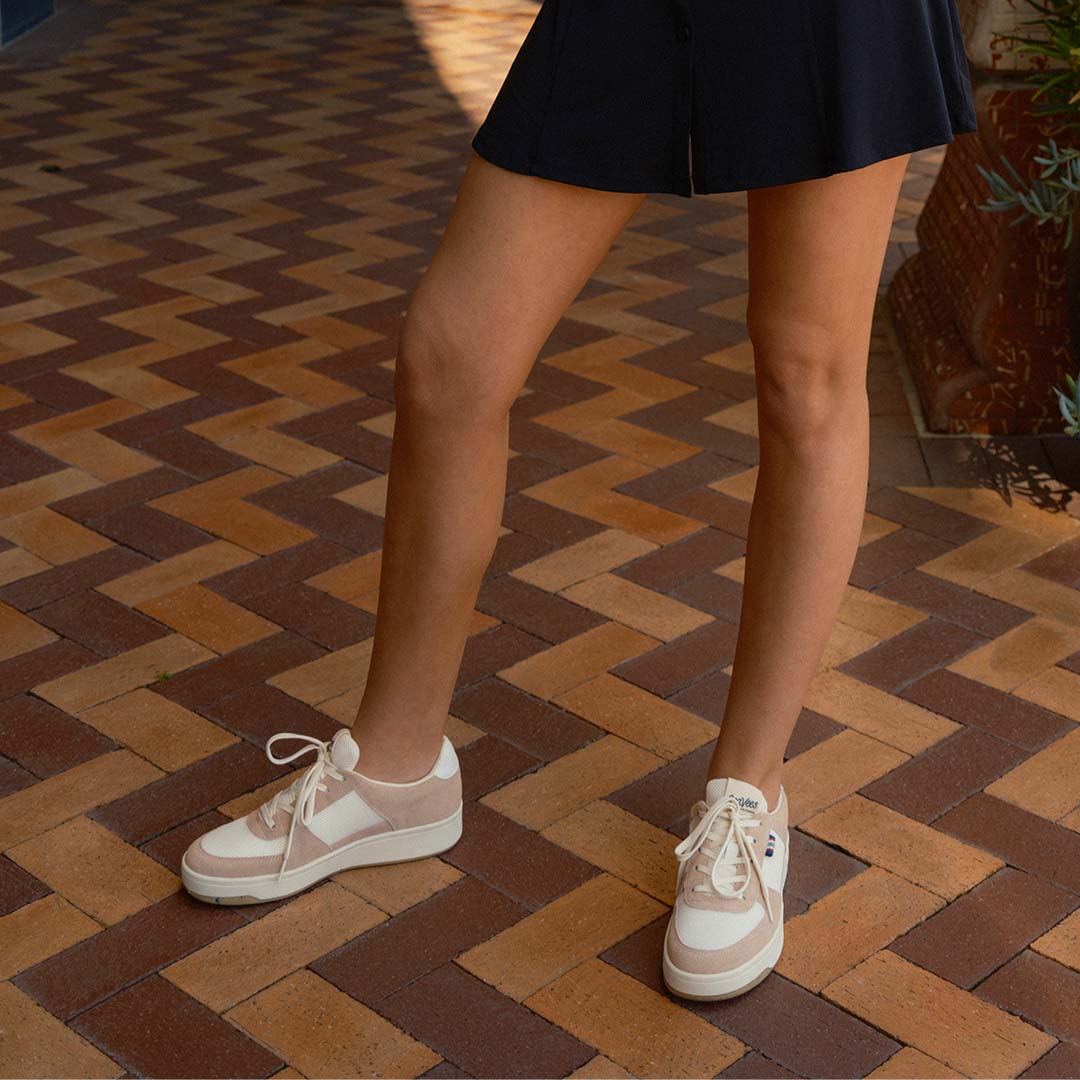 Person standing on a brick floor wearing black shorts and the Cardinal Sneaker in Pink Sand