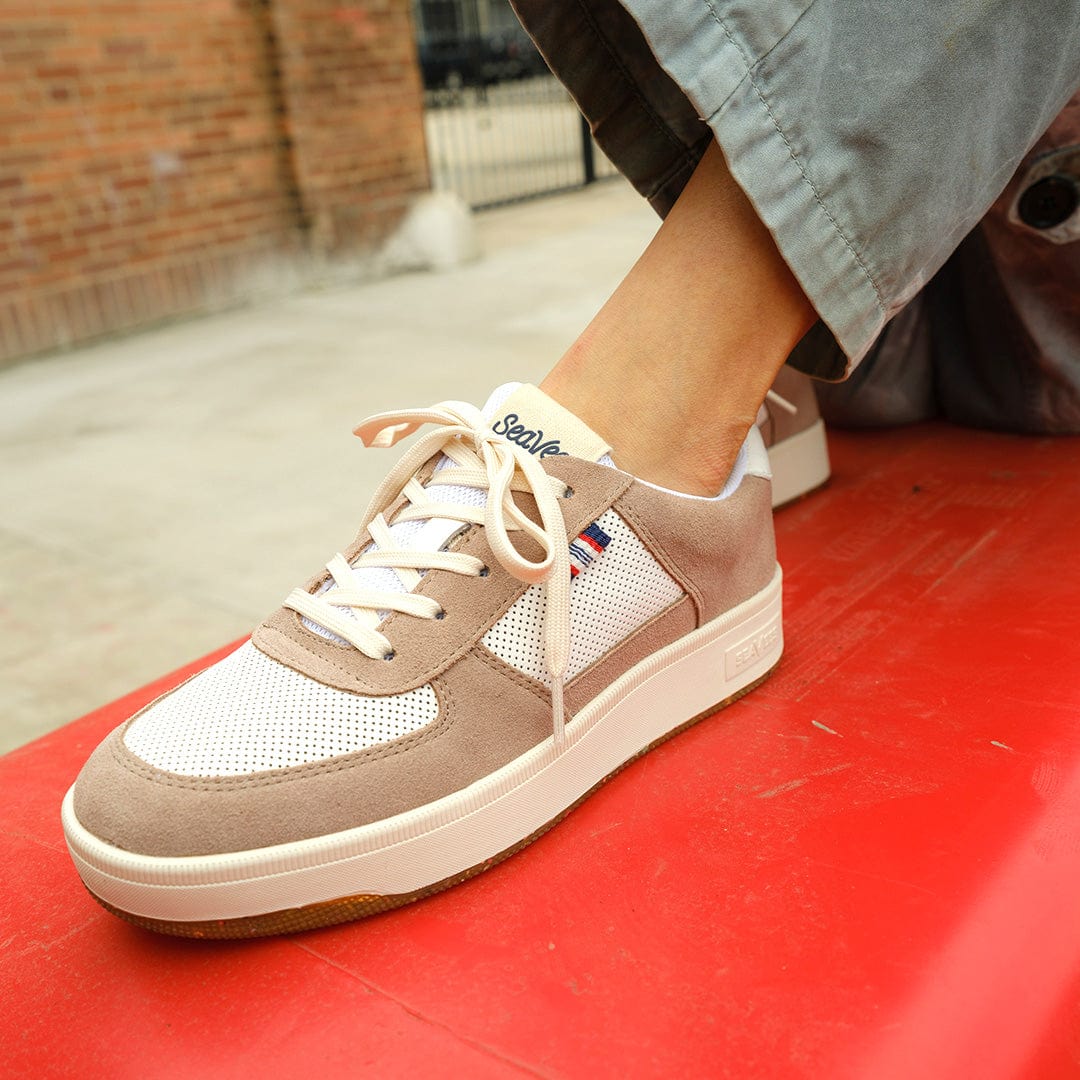 Close up view of someone sitting on a red bench with gray pants and the Cardinal Snekaer in Latte, showcasing the vintage-style SeaVees branding on the tongue and mesh detailing