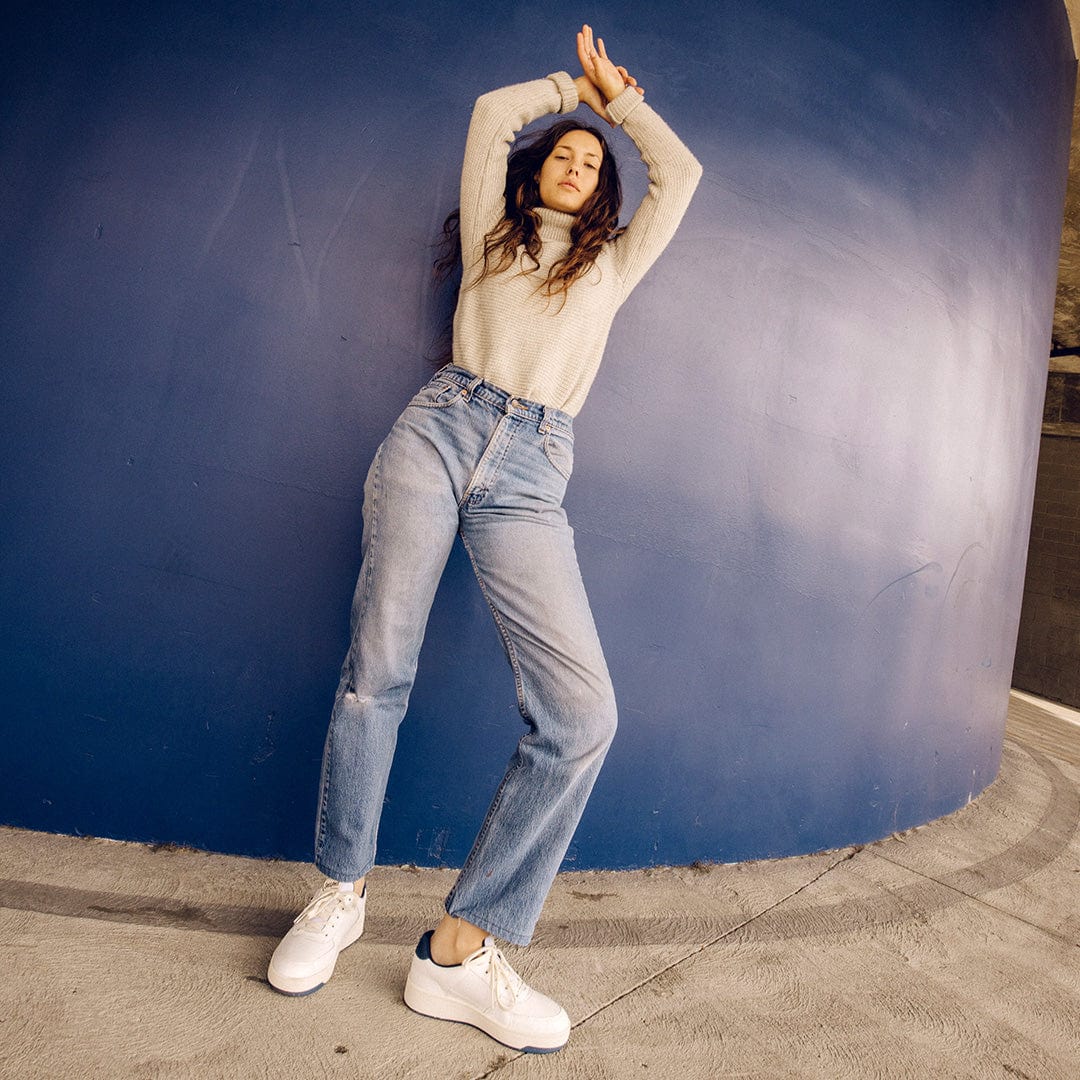 Person standing with arms over their head against a blue curved wall wearing a beige sweater and blue jeans with the Cardinal Sneaker in White Navy.