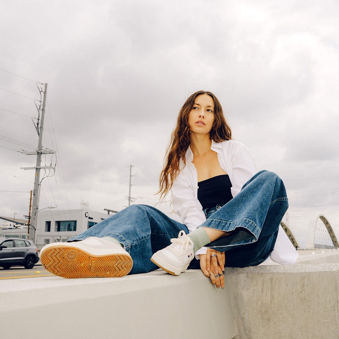 View from below of a person sitting on a concrete wall wearing a white and black top with blue jeans and the Cardinal Sneaker in white, showing off the colorful recycled sole