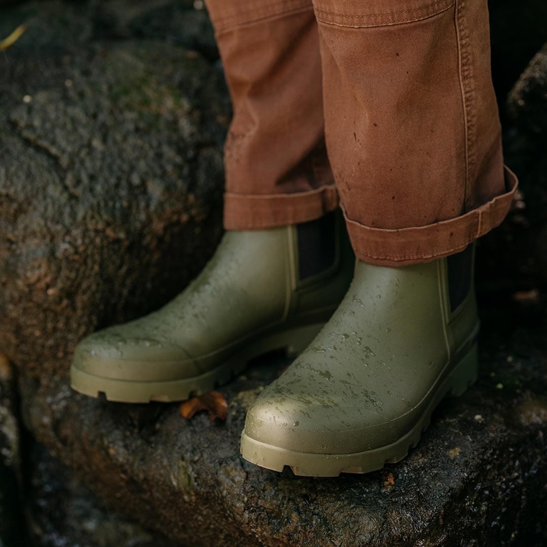 Feet in Bolinas Off Shore Boots in Military Olive, standing on a mossy rock outdoors.