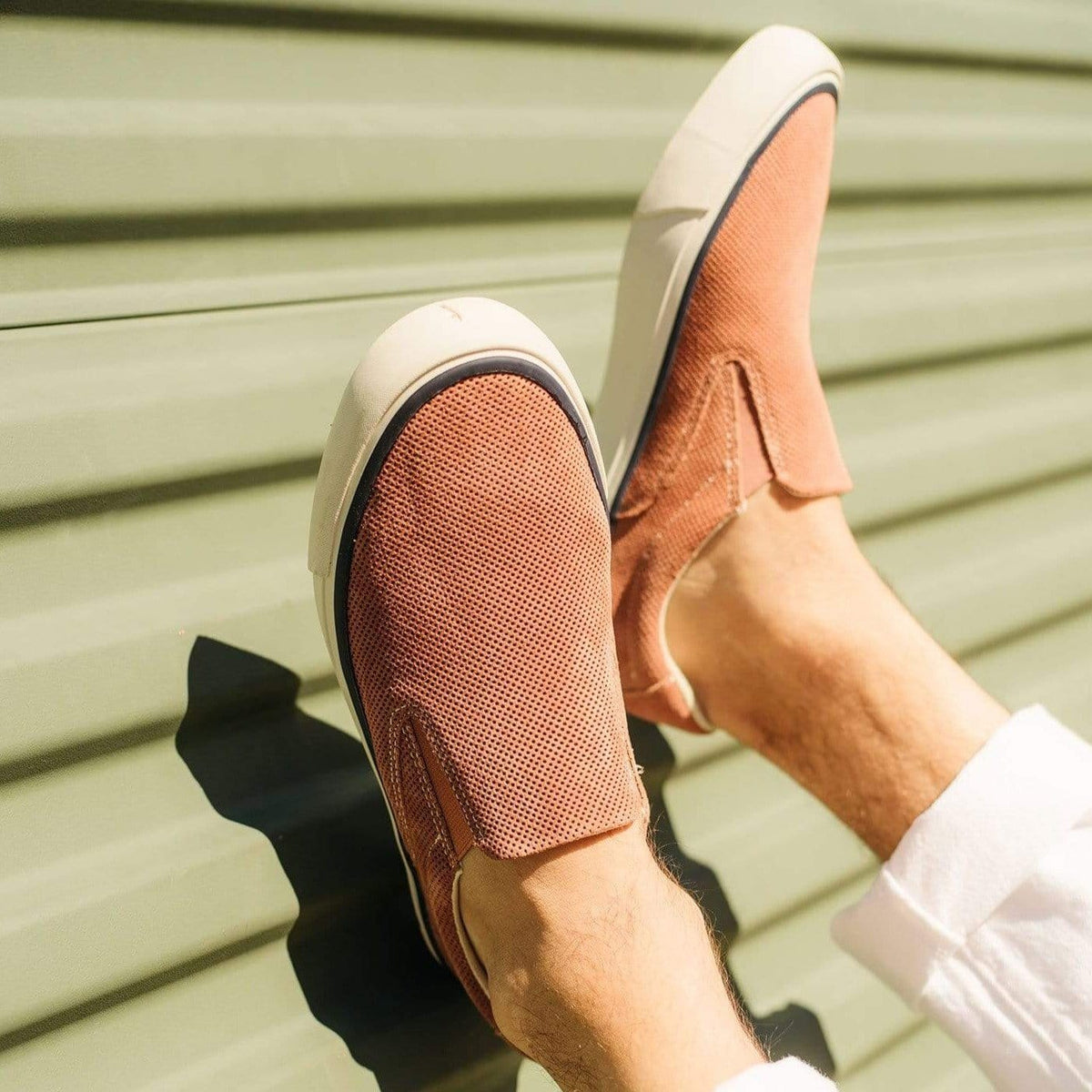 Men's Hawthorne Slip On in Terracotta suede, worn with white pants, feet raised against green siding