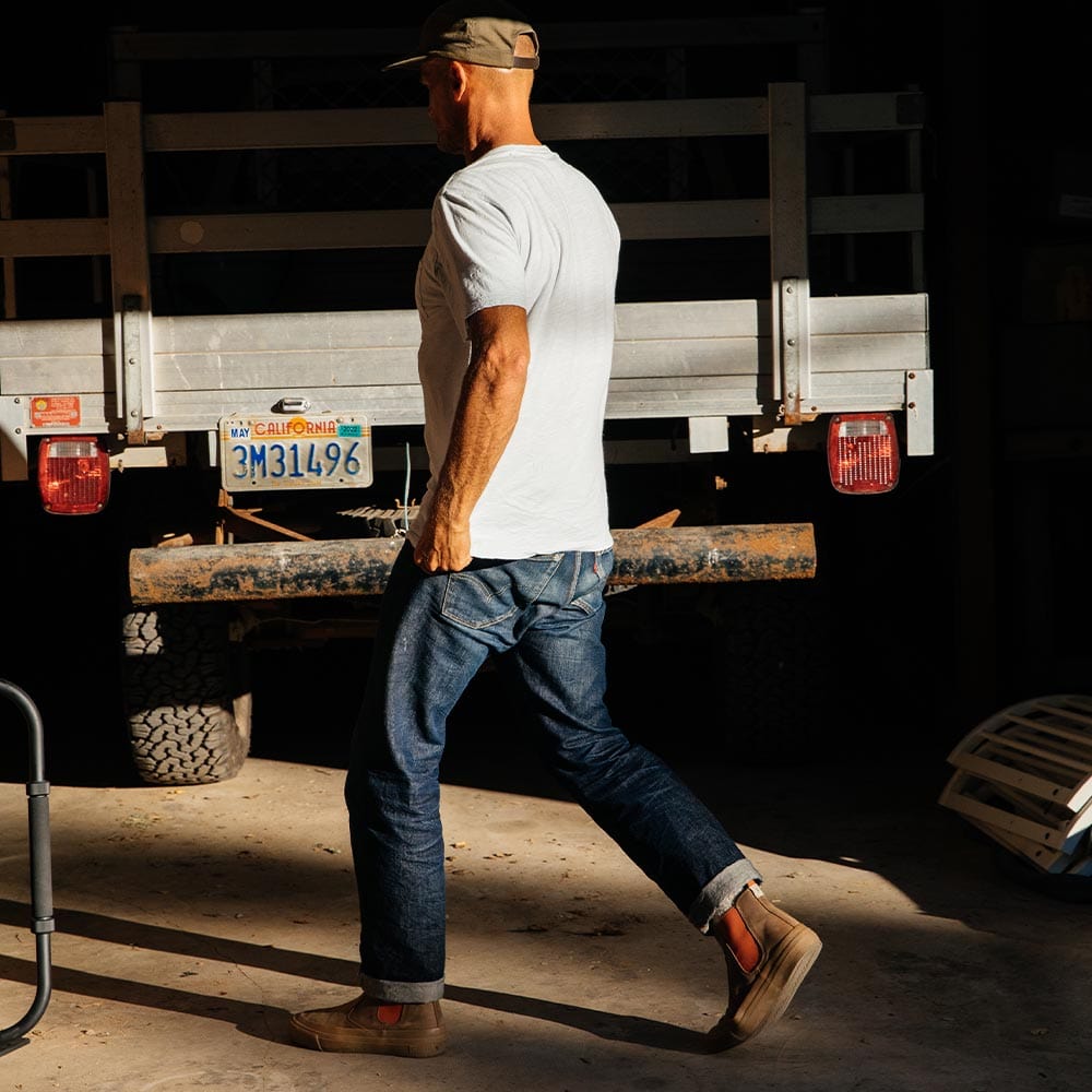 Man walking viewed from the side, wearing Elmwood Beyond & Back Boots, jeans, a T-shirt, and a cap, with a workshop in the background