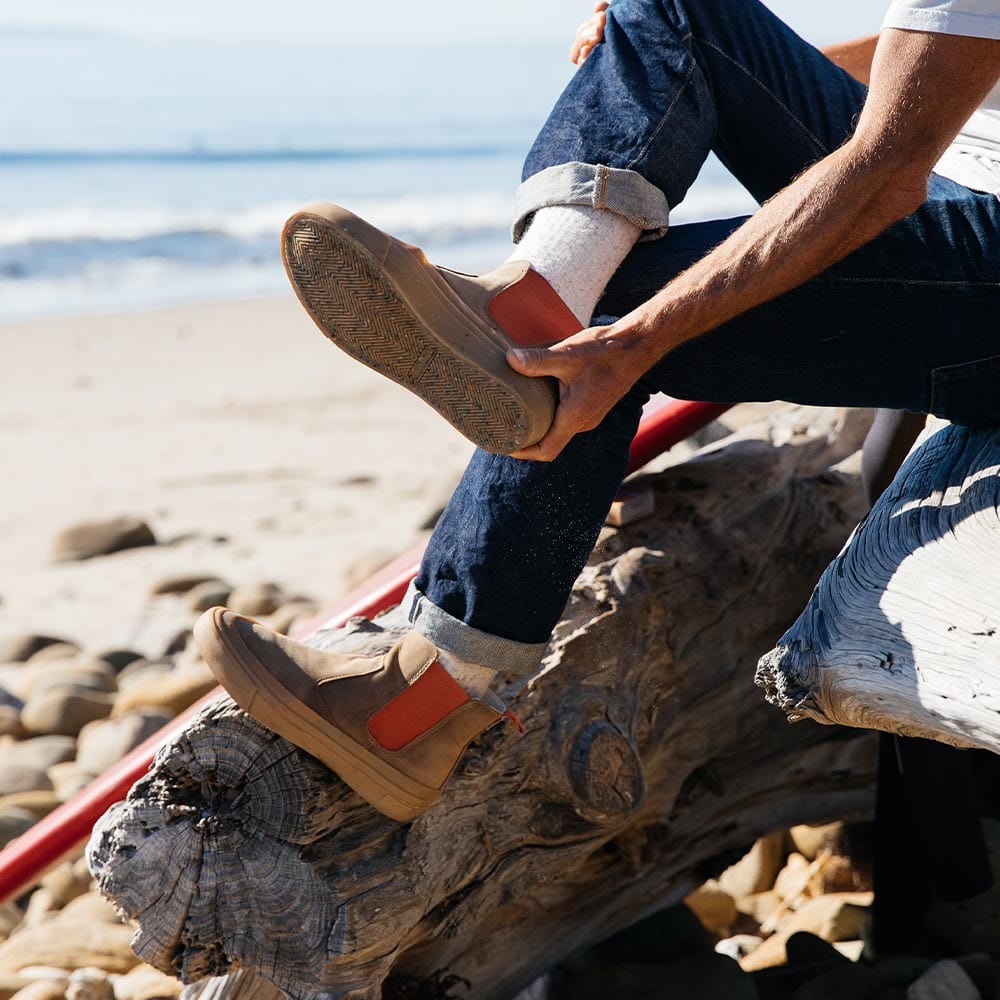 Close-up of Beyond & Back Boot in Elmwood, worn with cuffed jeans on a beach log, showcasing the boot's sole and side profile