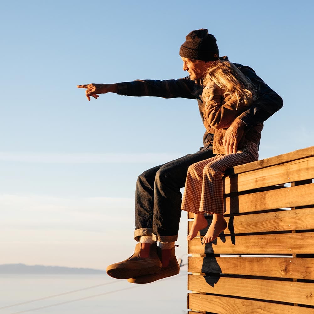 Person seated on wooden ledge wearing Beyond & Back Boots in Elmwood, pointing towards the horizon, with a clear sky background
