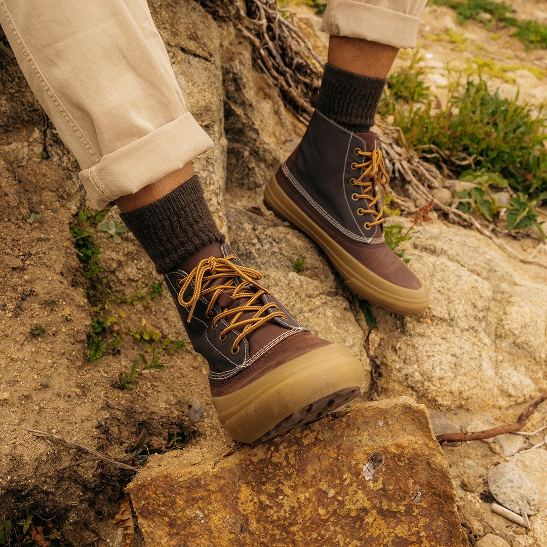 Person wearing Hickory/Charcoal Cascade Range Boots leaning against a white fence, highlighting their rugged outdoor style.