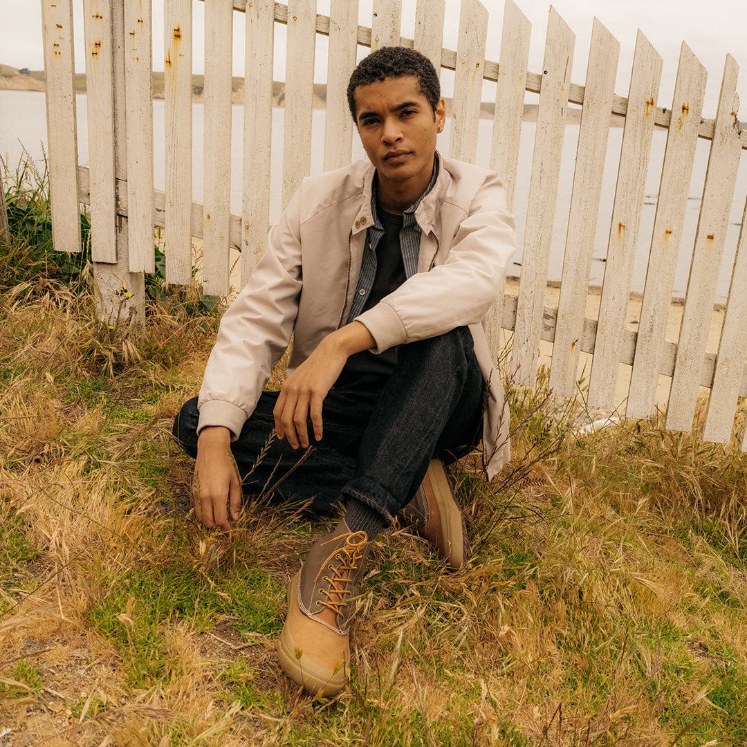Man sitting by a white fence in a natural setting, wearing Cascade Range boots in Cashew/Olive with rolled-up denim jeans.
