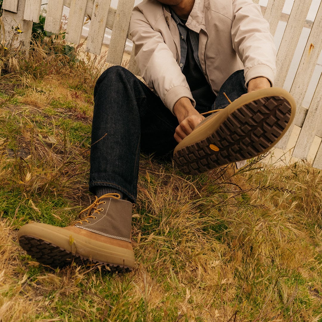 Relaxed pose of a man sitting on grass, showing the sole of Cascade Range boots in Cashew/Olive, demonstrating the grip.