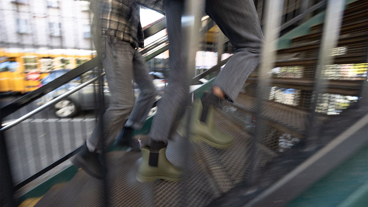 Load video: this is a video of two young people running around New York City wearing SeaVees new rainboots called The Bolinas Boot. They go on a train, step in puddles, and walk the streets of New York in comfort and style.
