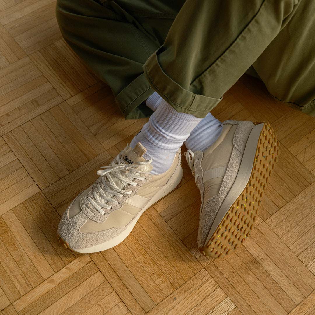 Up close lifestyle shot of a person wearing the Acorn Trainer in the color Cloud sitting on a hardwood floor.