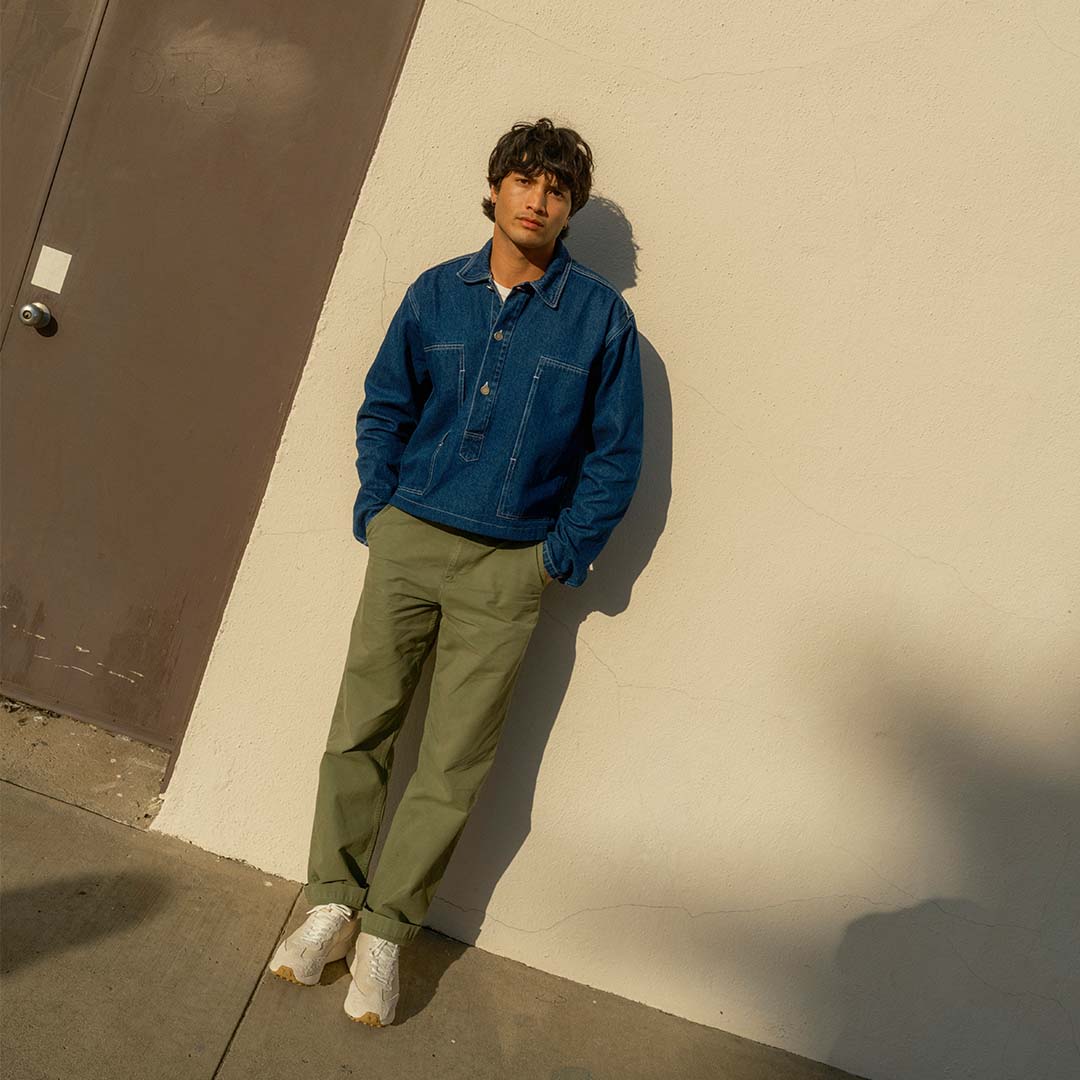 Person wearing the Acorn Trainer in the color Cloud along with a jean jacket and olive green pants standing on the sidewalk against a white wall.