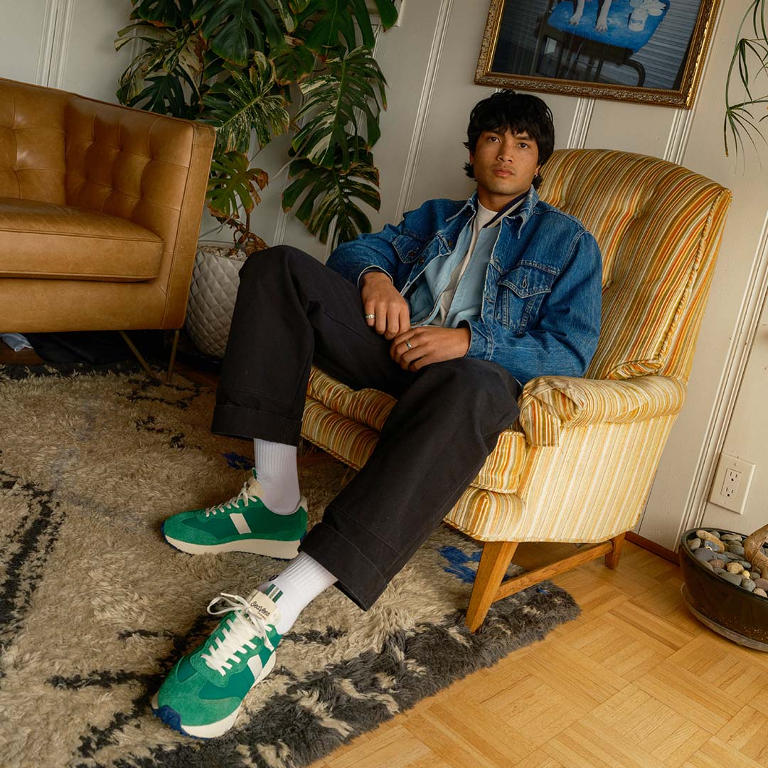 A full body shot of a person sitting in a chair with their left leg out showing off the Acorn sneaker in Grass Green.