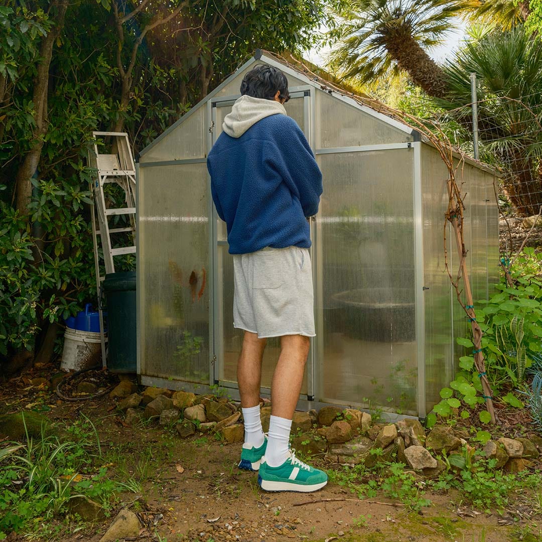 A rear view of a person standing in a garden, showcasing the back of the SeaVees 'Acorn' sneakers in Grass Green.