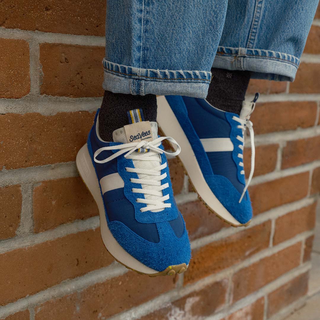 Close up view of someone sitting over a brick wall wearing jeans and the Acorn Trainer in Varsity Blue.