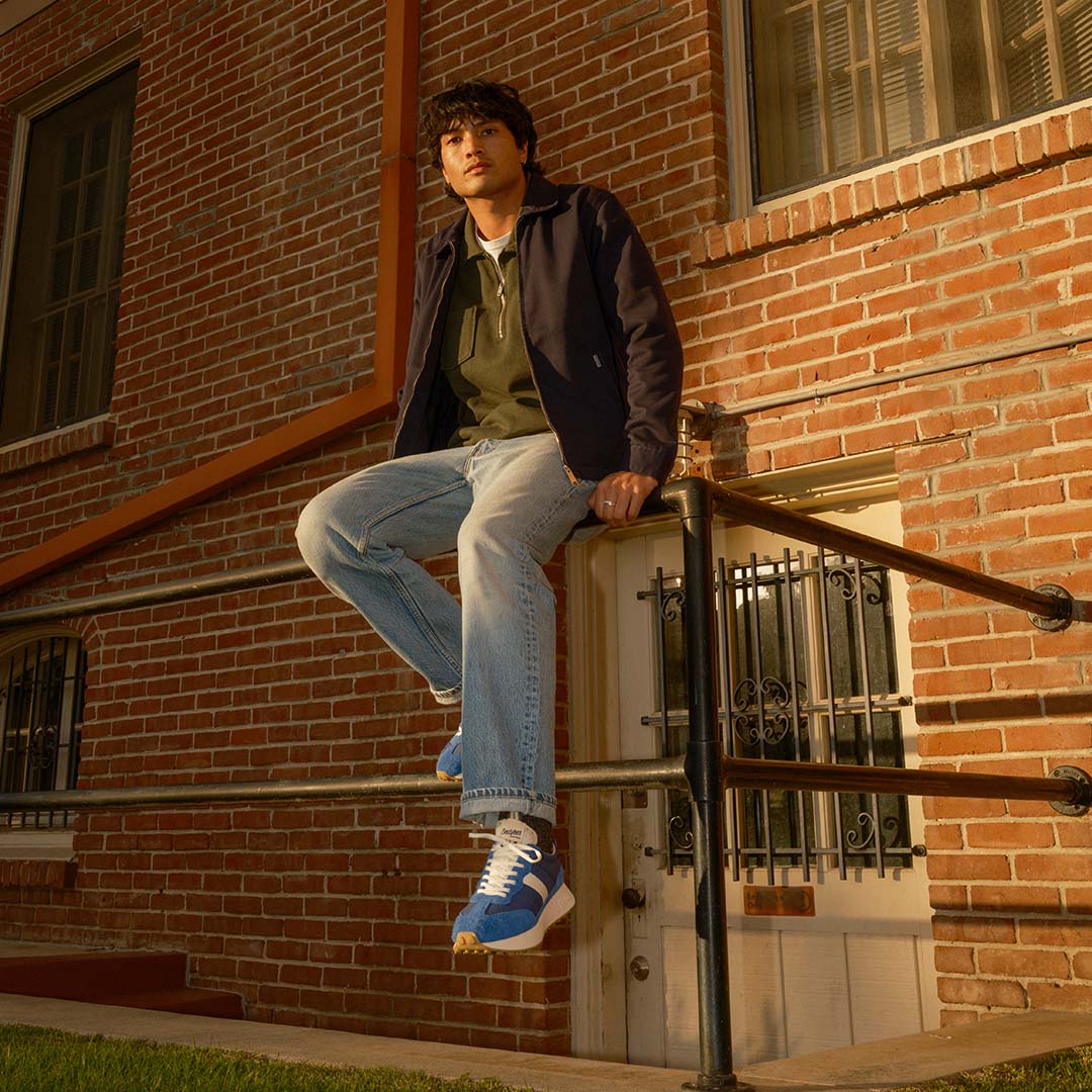 Person sitting on top of a railing wearing blue jeans and the Acorn Trainer in Varsity Blue.