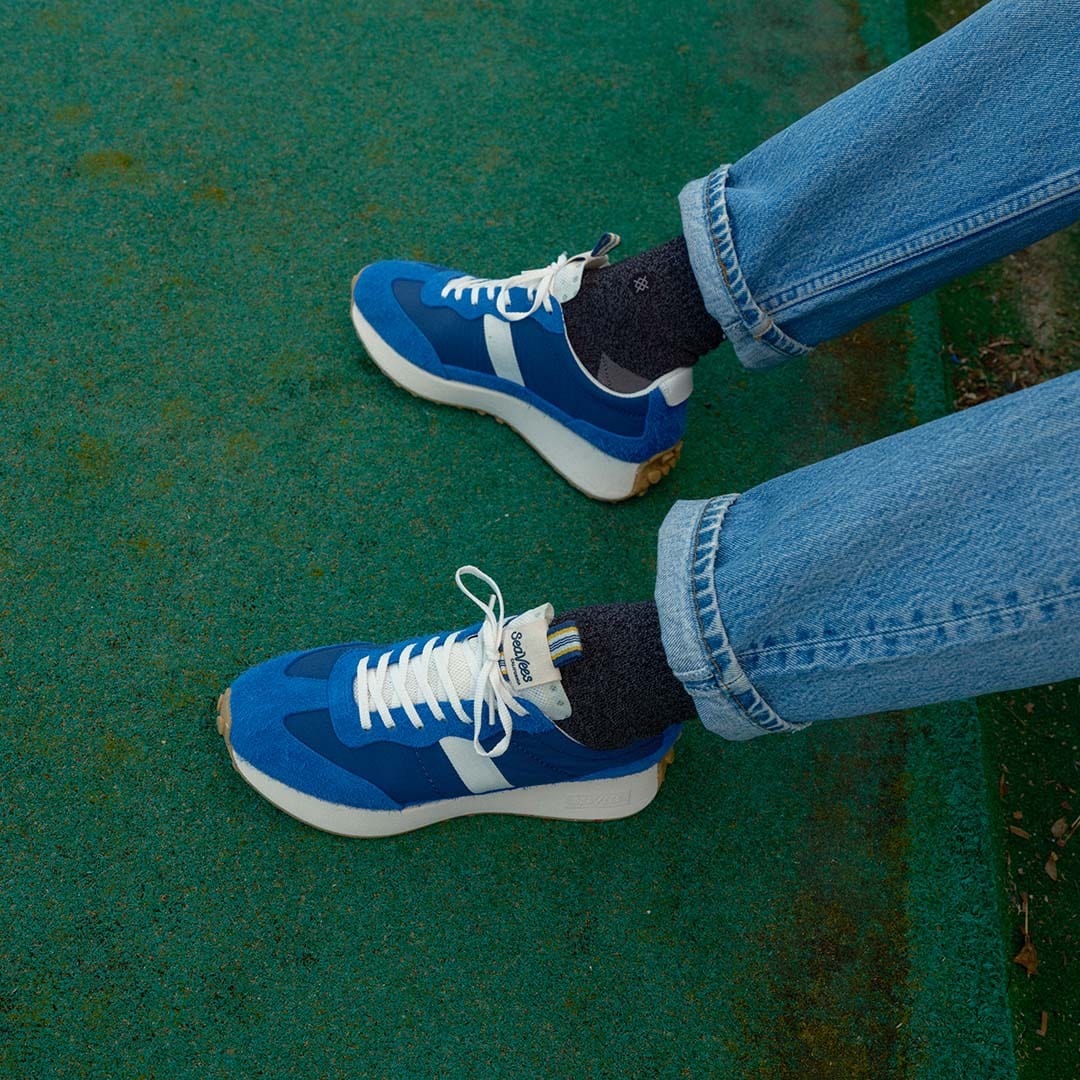 Close up photo of person wearing blue jeans, black socks, and the Acorn trainer in Varsity Blue while standing on a putting green.