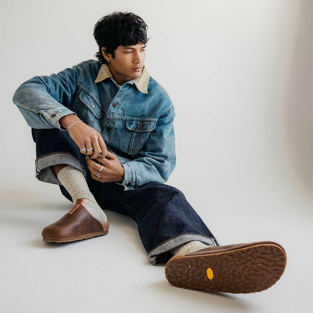 Men's Tiburon Trekker sandal in Elmwood leather, worn by a person seated on a white floor in a denim jacket