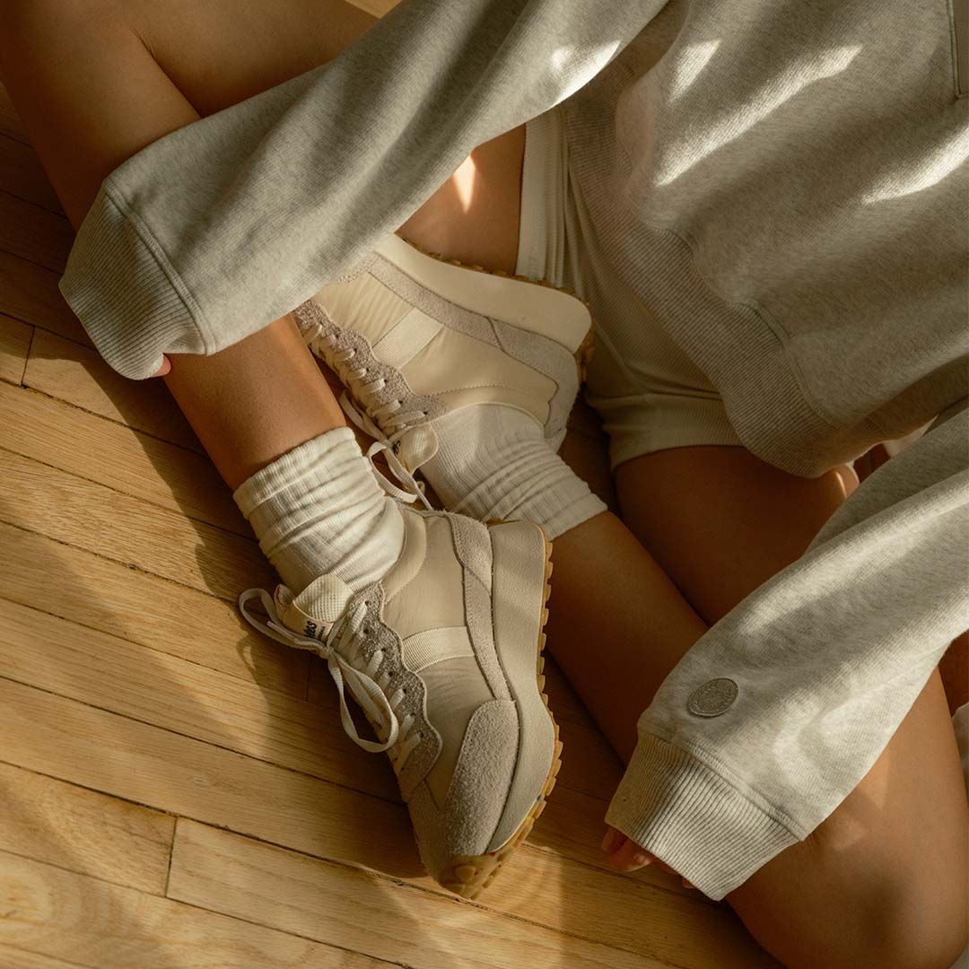 Close up shot of a person sitting cross-legged wearing the Acorn Trainer in the color Cloud on a hardwood floor.