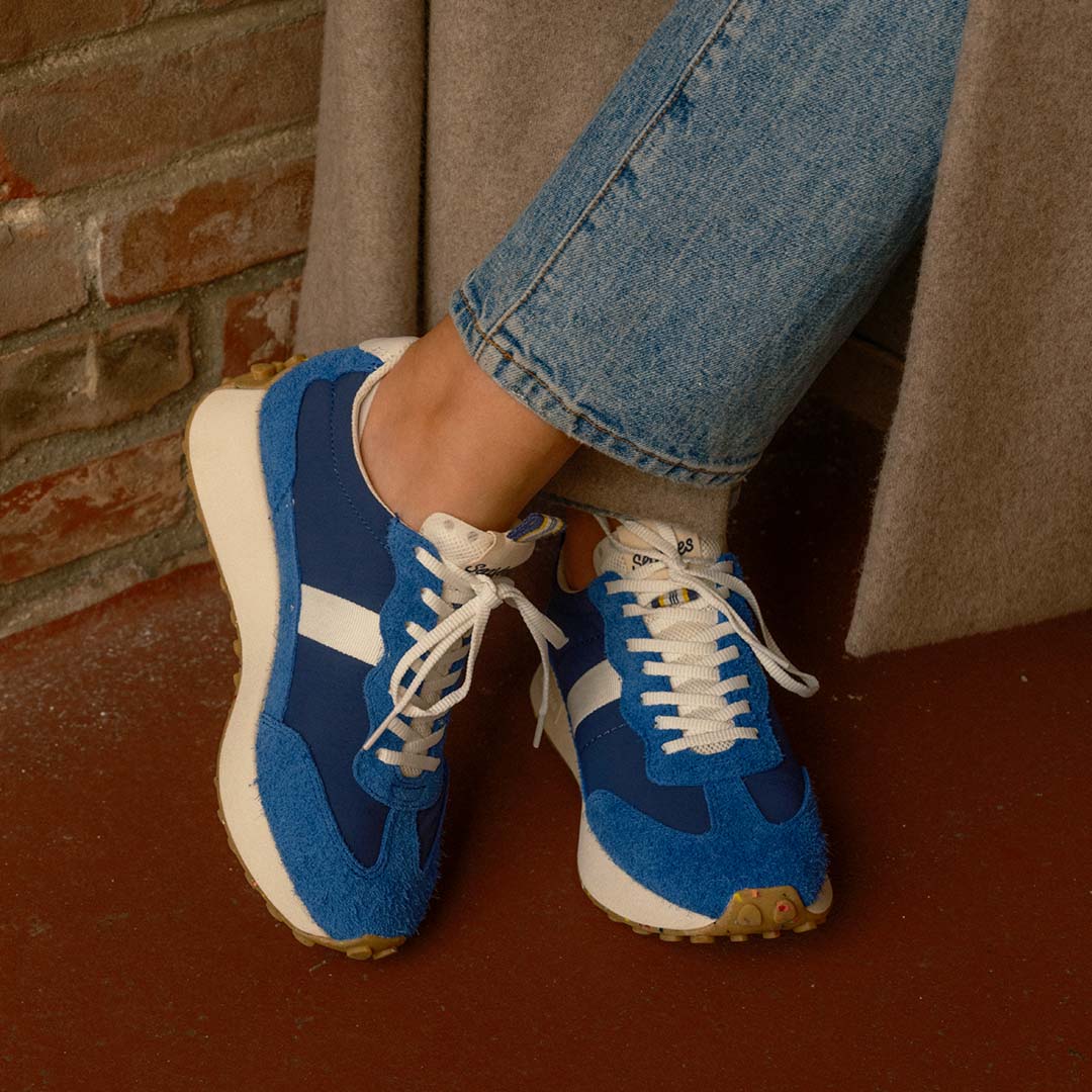 Closeup of a person wearing the Acorn Trainer in Varsity Blue standing against a brick wall wearing jeans.