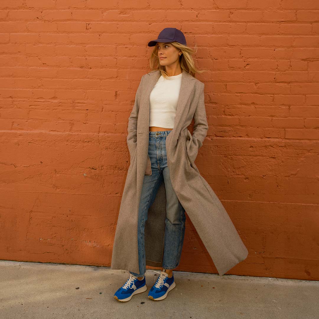 Person wearing the Acorn Trainer in Varsity Blue and standing against a brick wall, styled in jeans, a white tee shirt, and trench coat.