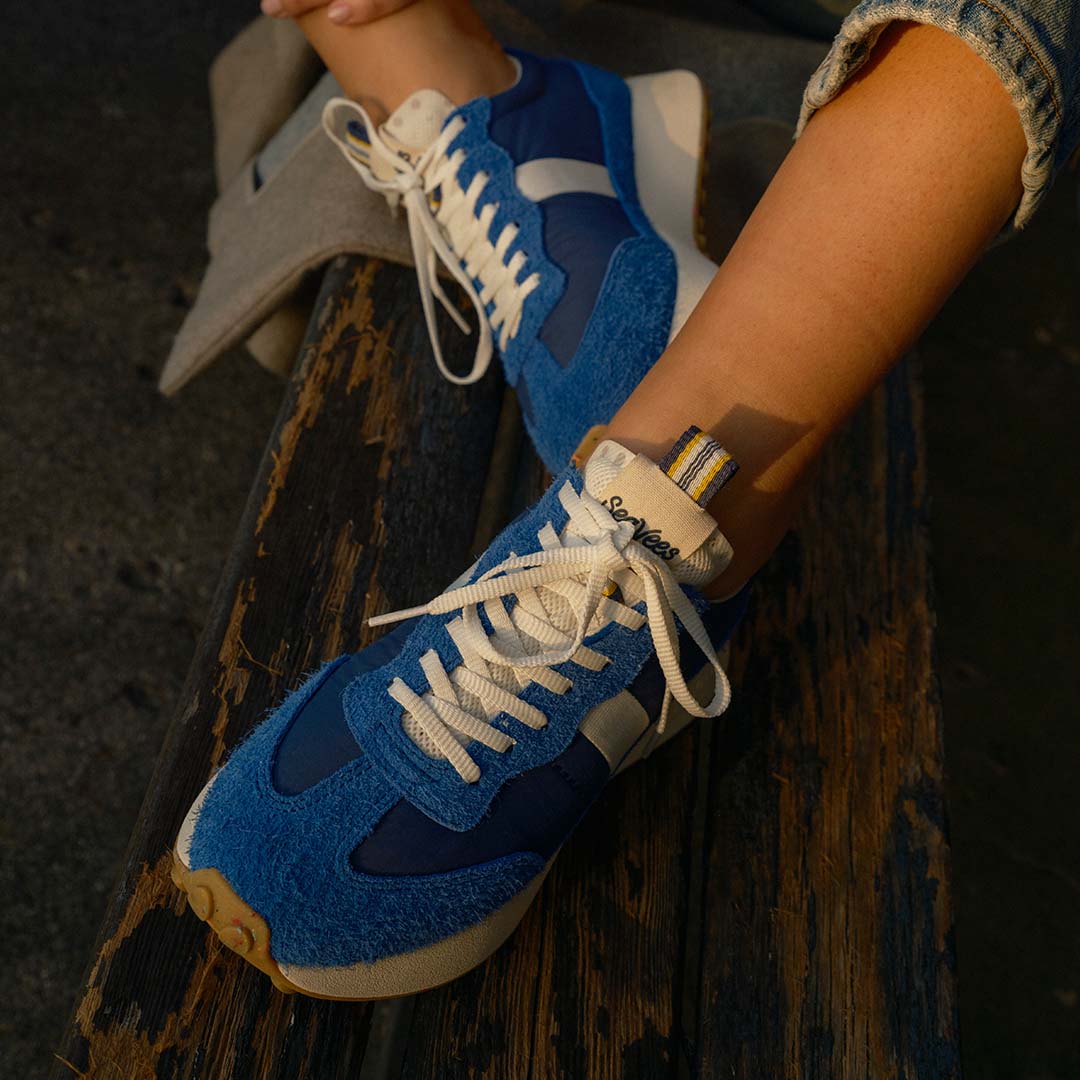 Close up view of a person sitting on a wooden bench wearing the Acorn Trainer in the Varsity Blue color, showing the detail of the laces and pull tab.