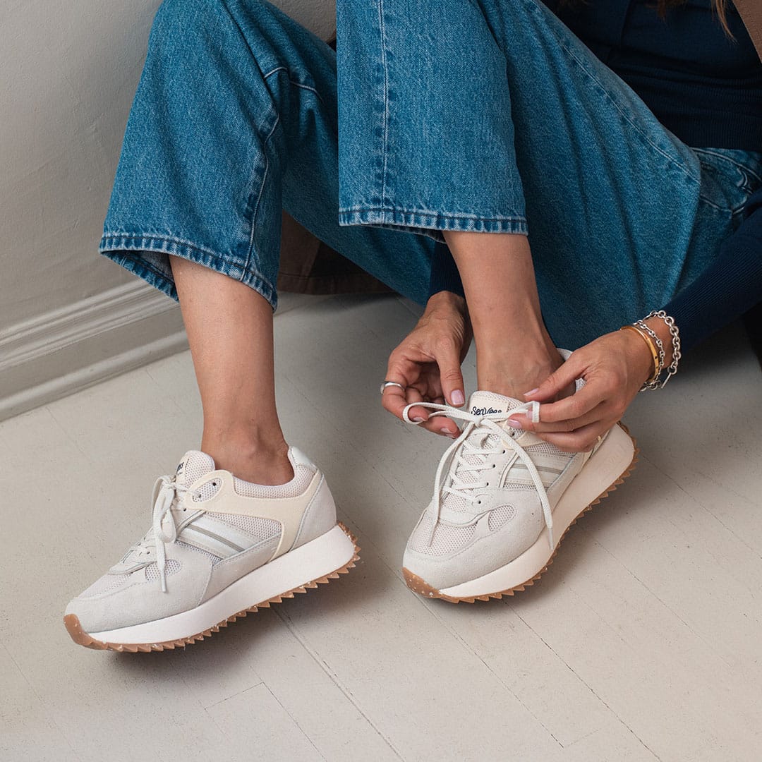 Women's Causeway Trainer sneaker in Cloud mesh and leather, worn with jeans, laces being tied on a white floor