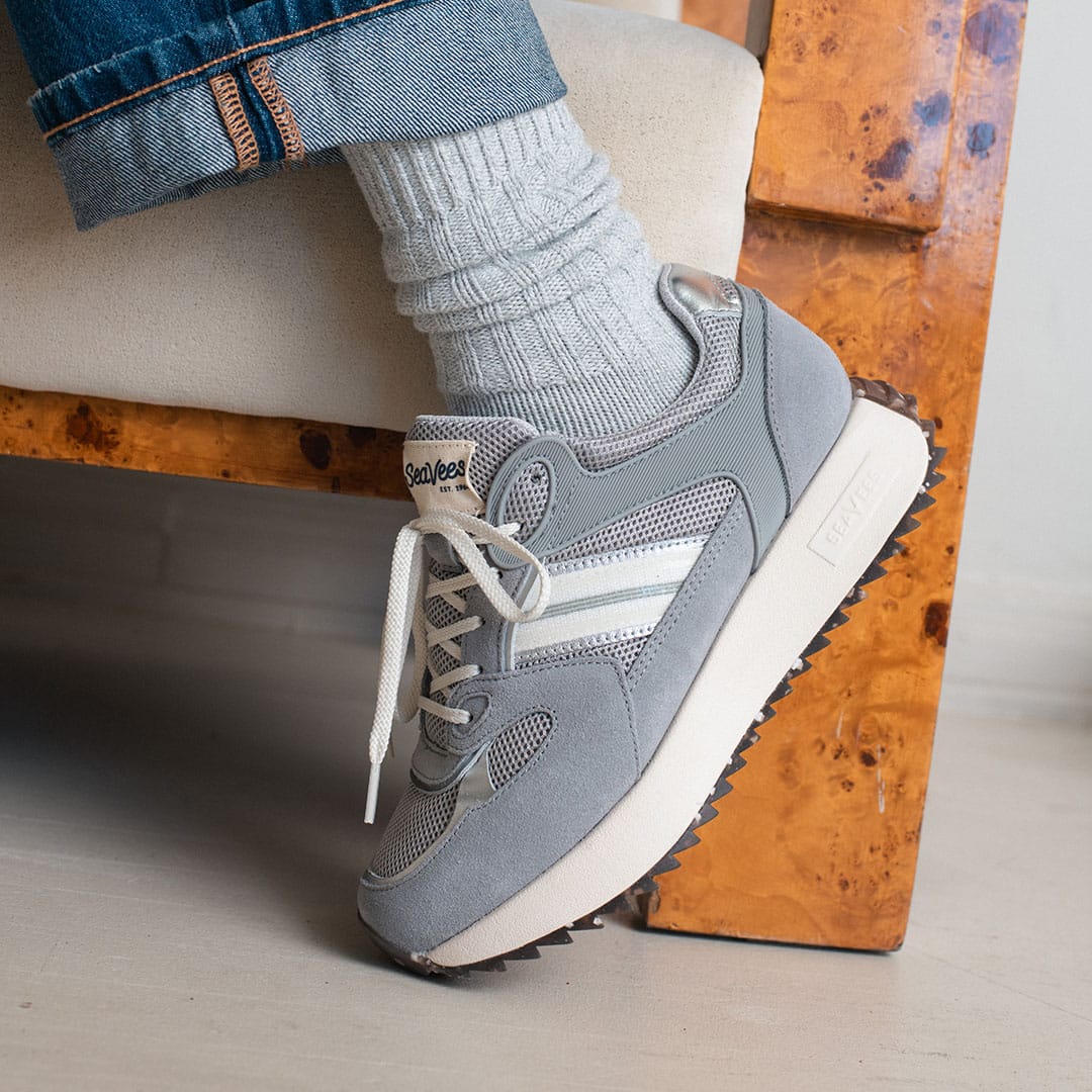 Women's Causeway Trainer sneaker in Moon Rock mesh and leather, worn with gray socks, seated beside a wooden chair
