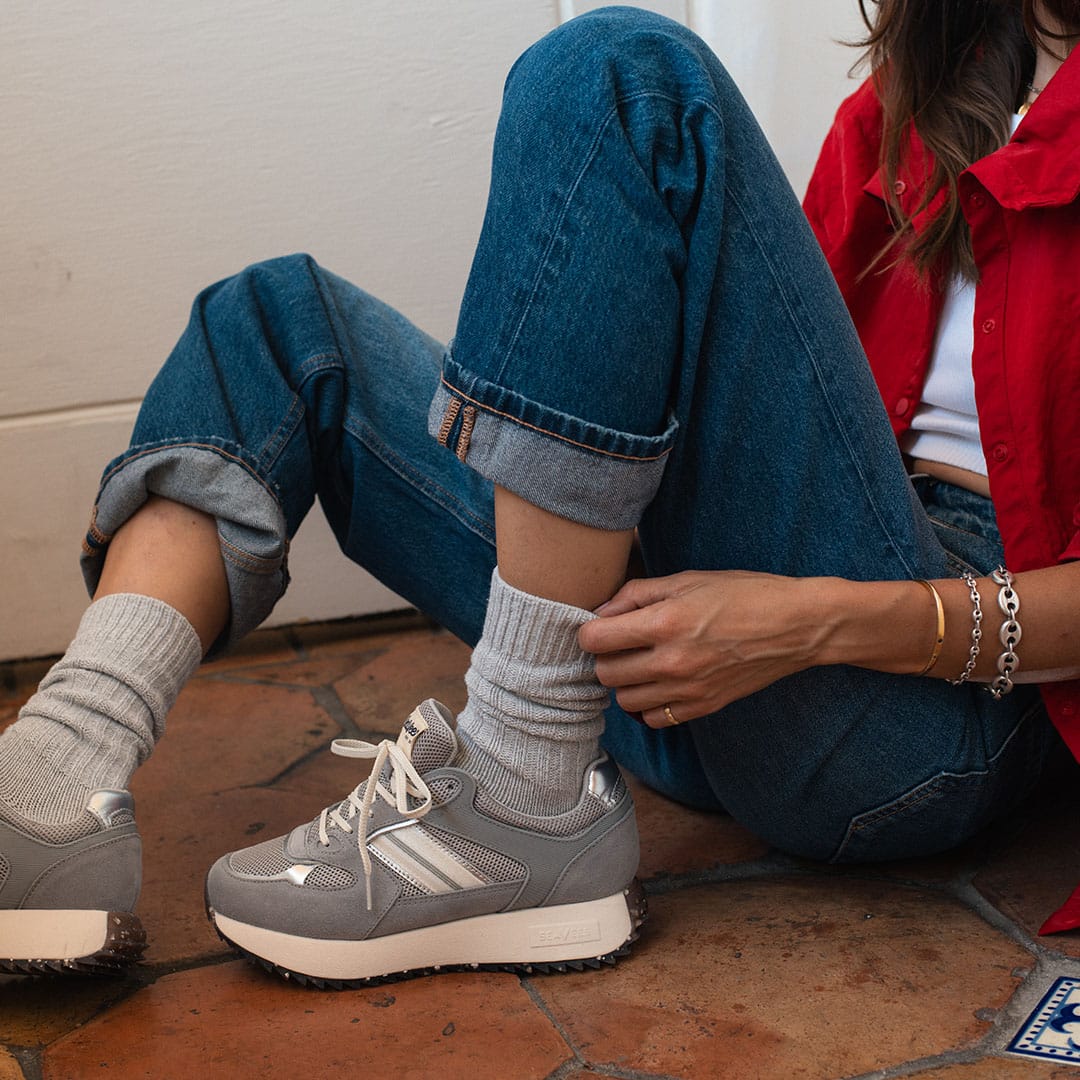 Women's Causeway Trainer sneaker in Moon Rock mesh and leather, worn with gray socks and rolled jeans, seated on tile
