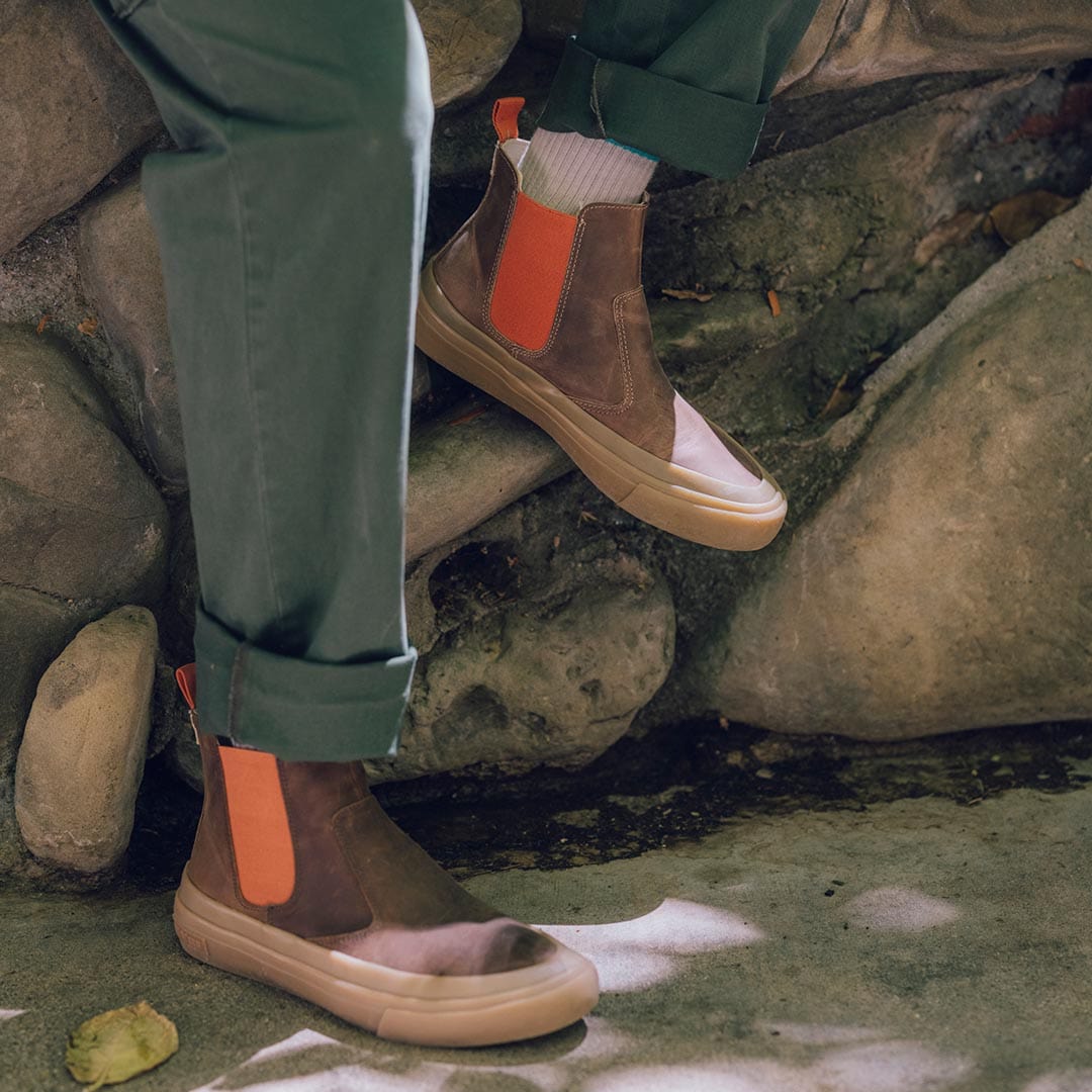 Beyond & Back Boots in Elmwood on feet, crossed at the ankles, against a natural stone backdrop with green foliage