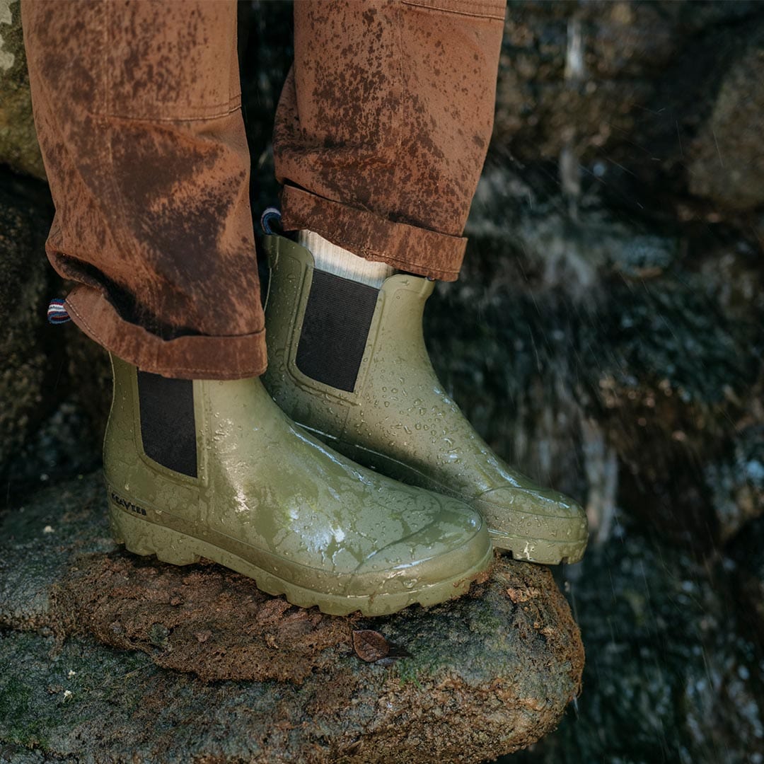 Bolinas Off Shore Boots in Military Olive worn outdoors, splattered with mud on a rocky ledge.