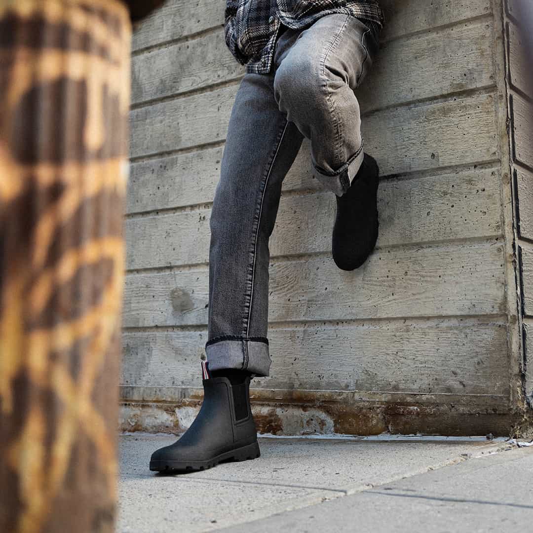 Close-up of a person resting their foot against a brick wall, showcasing the Bolinas Off Shore Boot in Black with visible heel loop