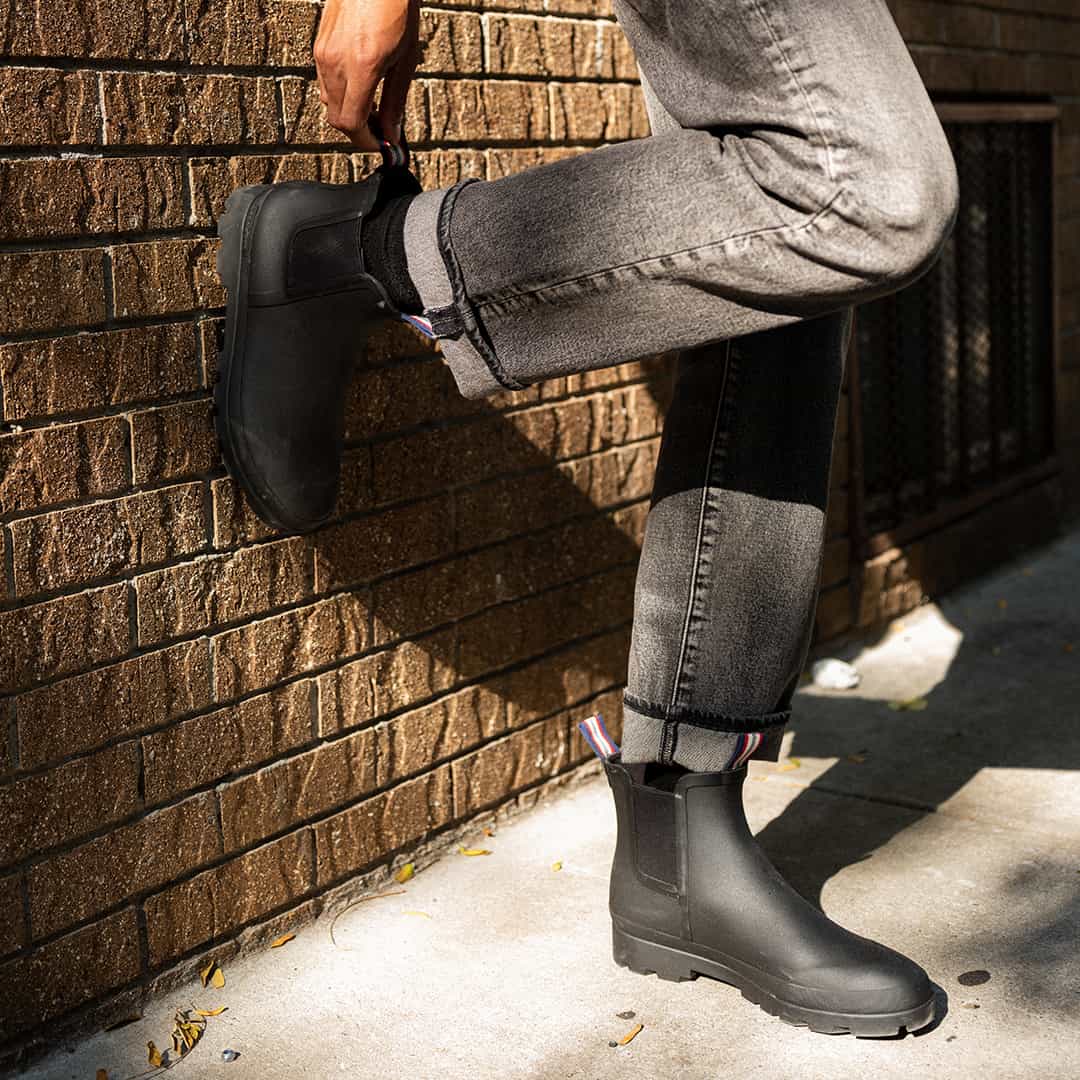 Close-up of a person resting their foot against a brick wall, showcasing the Bolinas Off Shore Boot in Black with visible heel loop