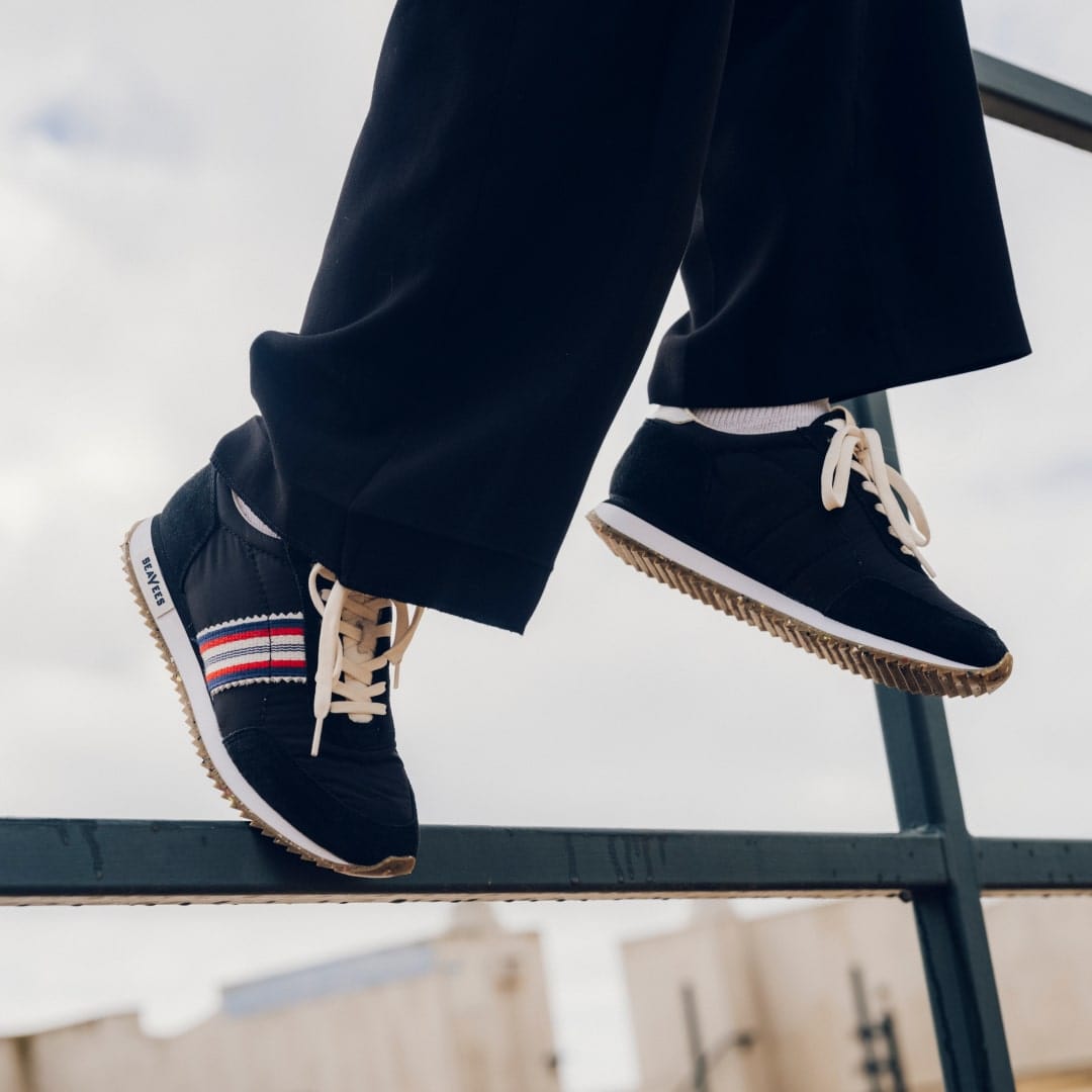 Close up view of someone sitting on a railing wearing dark colored pants and the Royal Runner sneaker in Black